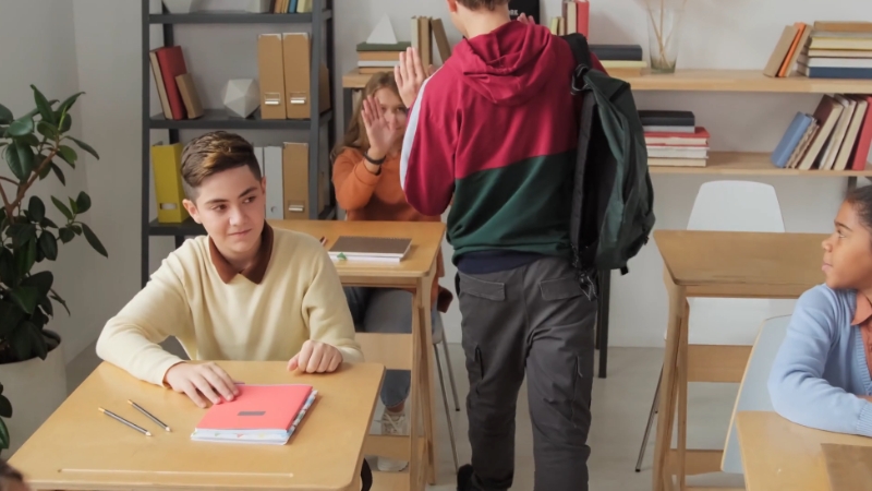 Students interact casually in a classroom with desks, bookshelves, and school supplies in view
