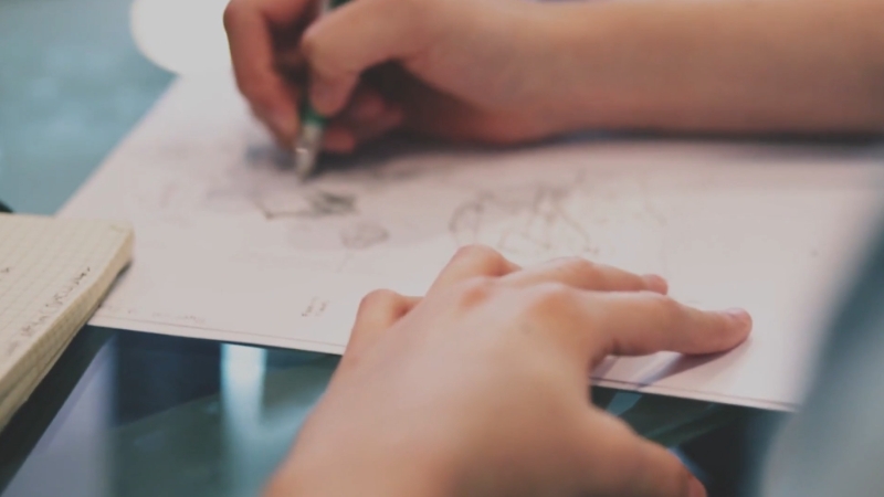 A student sketches on paper during class while another hand rests nearby on the desk