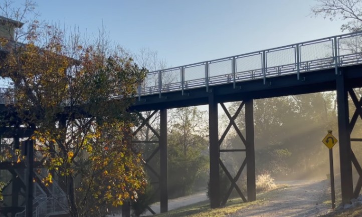 A bridge spans a river, with a walking path alongside it, surrounded by greenery and blue sky