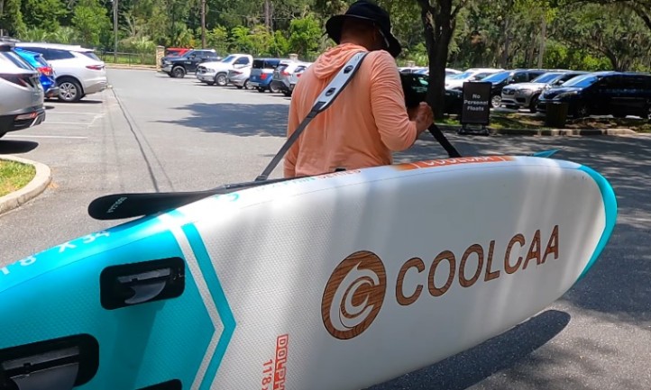 Man carrying a large, inflatable COOLCAA stand-up paddleboard with a shoulder strap in a sunny parking lot