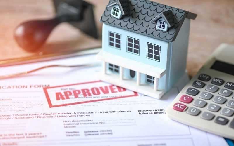 Model house on a home improvement application stamped approved, with a calculator and paperwork